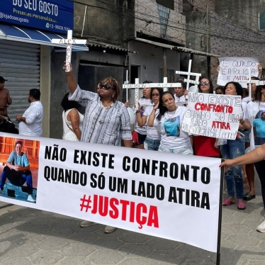  The image shows a group of people participating in a protest. The protesters hold banners and wear t-shirts with a person's face. One prominent banner read, in Portuguese, "There is no confrontation when only one side shoots" with the word "JUSTICE" emphasized below. 