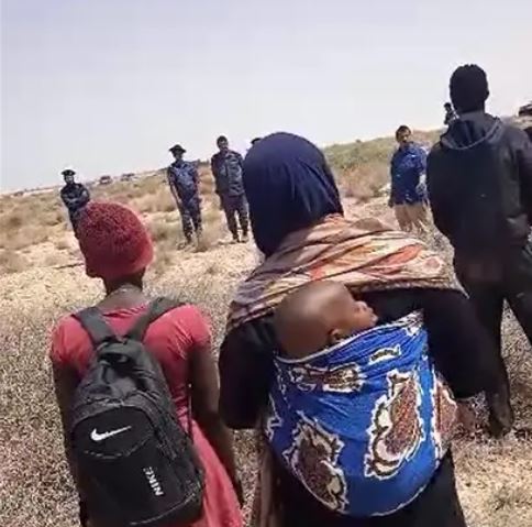 Un grupo de migrantes y solicitantes de asilo negros de múltiples nacionalidades africanas, entre ellos una mujer y su bebé, abandonados en el desierto durante días tras ser expulsados de Túnez, permanecen en la zona de seguridad de la frontera entre Túnez y Libia frente a un equipo de noticias de Al Jazeera y soldados libios, 11 de julio de 2023.