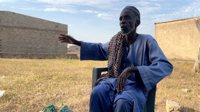 A man sits in front of a grassy area and gestures with his hand.