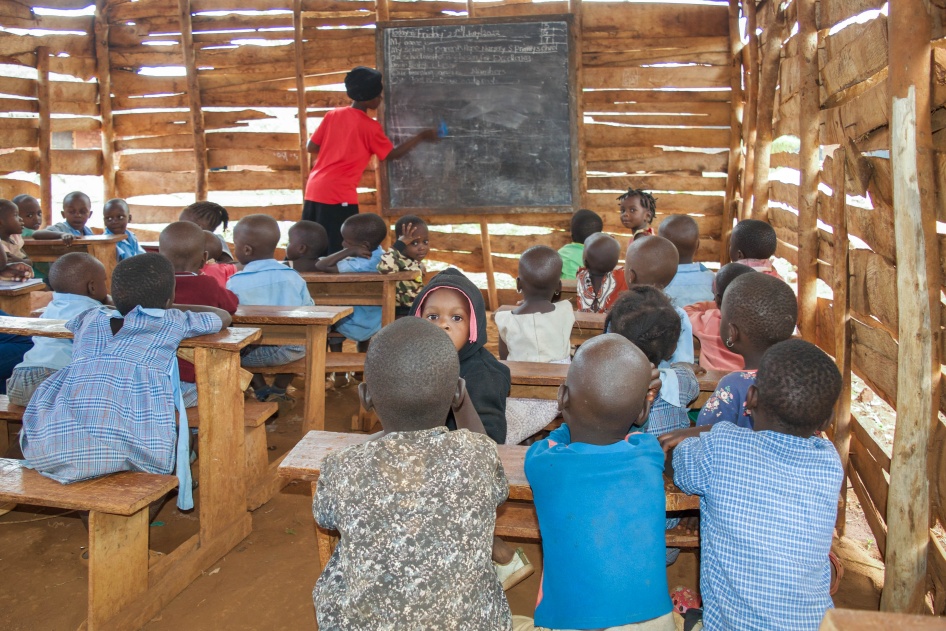 Young children sit in a rural classroom while a teacher writes on a blackboard